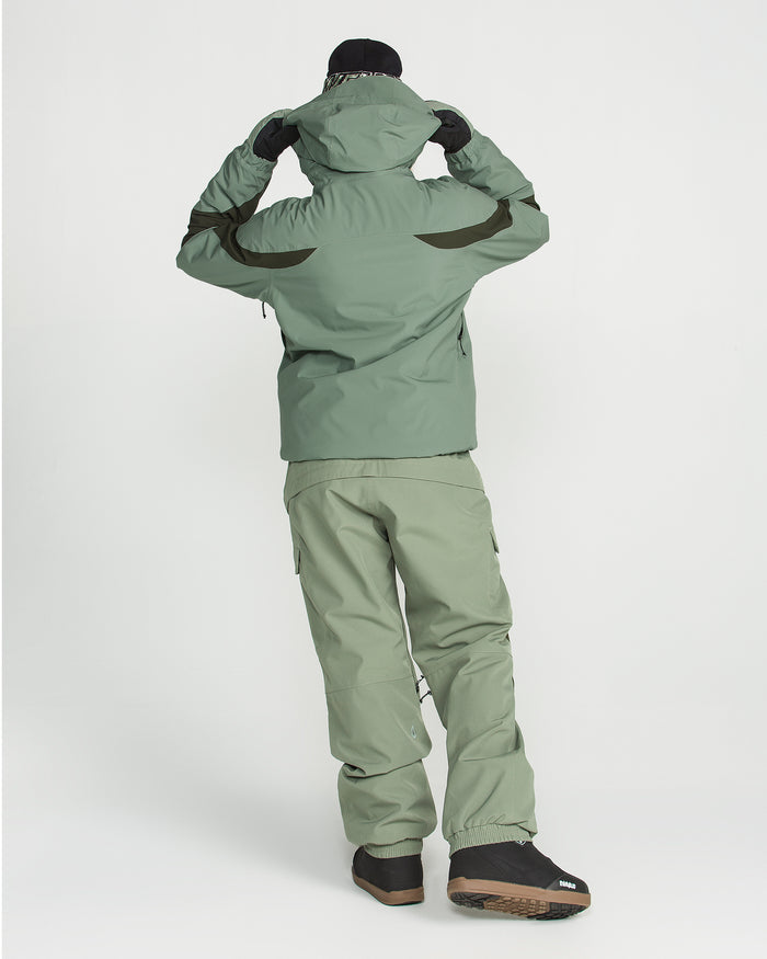 A person is seen from behind, adjusting their hood while wearing the Volcom Women’s Shelter 3D Stretch Jacket and matching pants in green. The outfit, highlighted by waterproof insulation and a PFAS-free DWR finish, is styled with black gloves, brown boots, and a black hat against a plain white background.