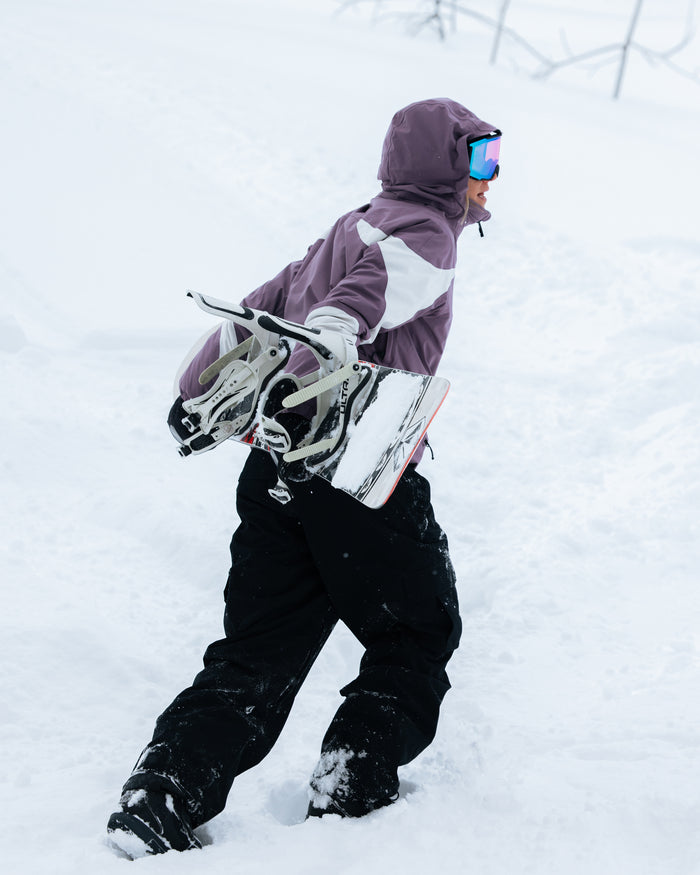 A snowboarder in a Volcom Women’s Shelter 3D Stretch Jacket in purple and black pants walks uphill in deep snow, carrying a snowboard under one arm.