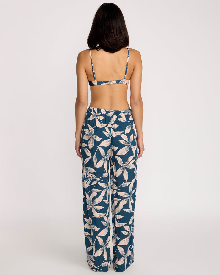 A woman faces away, wearing a strappy bikini top and Volcom Women's Shore Shot Pants in Harbor Blue with a blue and white leaf pattern. Her arms rest at her sides against a plain white background, showcasing chic beachwear style.