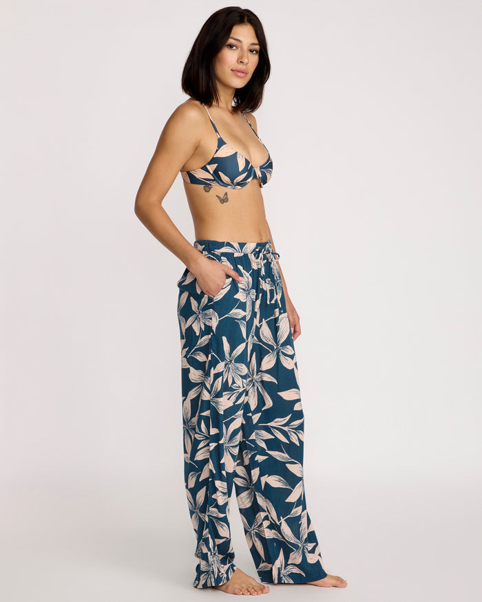 A woman with straight, shoulder-length dark hair and a butterfly tattoo on her side stands barefoot against a plain background, wearing a floral bikini top and Volcom Women's Shore Shot Pants in Harbor Blue with white leaf patterns.