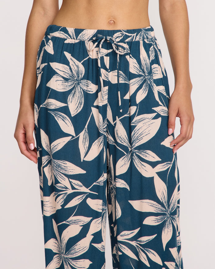 A person models Volcom Women's Shore Shot Pants in Harbor Blue, featuring a blue and white floral pattern, high waist, wide-leg fit, and drawstring waist, shown from midriff to mid-thigh.