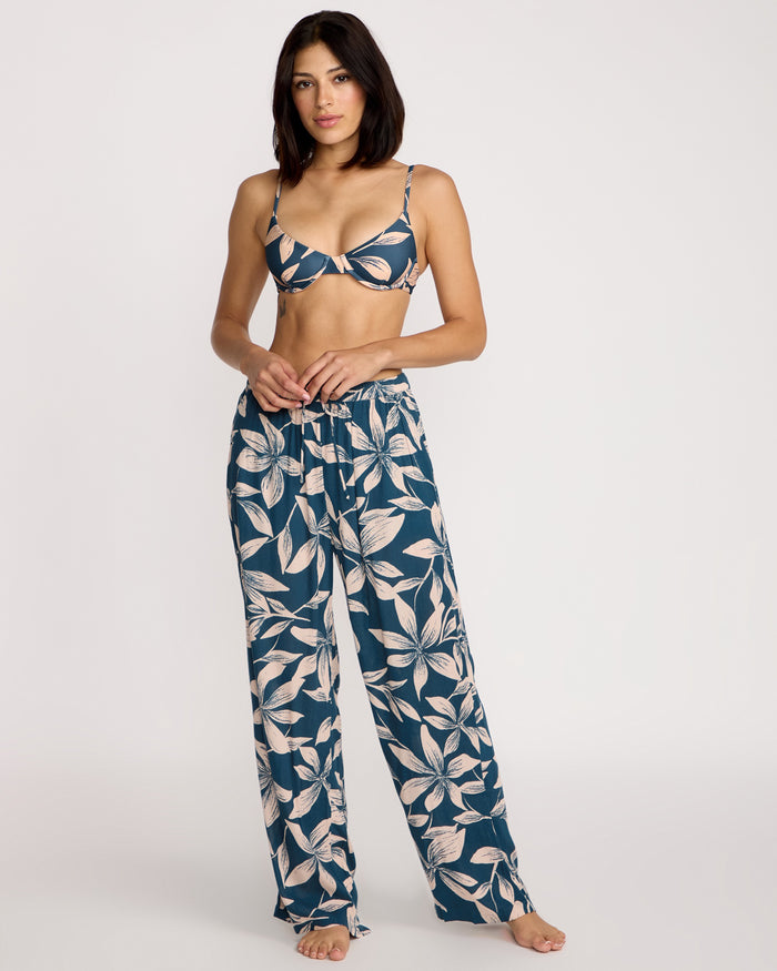 A woman stands barefoot in Volcom Women's Shore Shot Pants—Harbor Blue floral wide-leg beachwear—paired with a matching bralette top, posing against a plain white background.