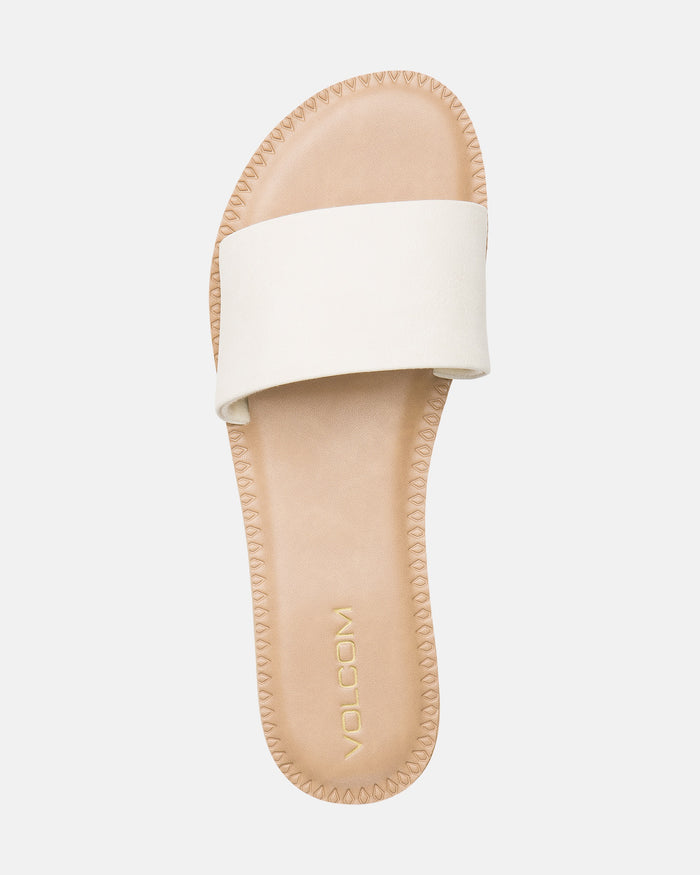 The Volcom Women's Simple Slide is a tan flat sandal featuring a wide white strap, visible stitching around the sole, and "VOLCOM" in gold lettering on the footbed.