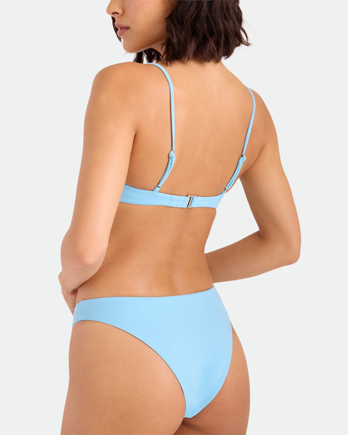 A woman with shoulder-length dark hair is seen from the back, wearing the Volcom Women's Simply Seamless Bralette in light blue, made from recycled nylon with adjustable straps and a clasped top, against a plain white background.