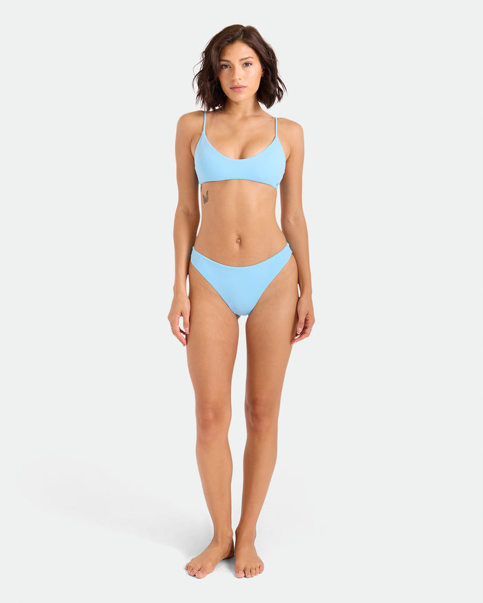 A woman with short brown hair stands barefoot against a white background, wearing the Volcom Women's Simply Seamless Bralette in light blue. She faces forward with a neutral expression, arms relaxed at her sides.