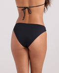 A woman with medium skin tone and long wavy hair is seen from the back wearing the Volcom Women's Simply Seamless Hipster Full black bikini bottom, highlighting its sleek fit against a simple backdrop.