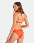 A woman with light skin and brown hair models the Volcom Women's Simply Seamless Hipster Full bikini in orange with adjustable straps, made from recycled nylon, shown from the back against a plain white background.