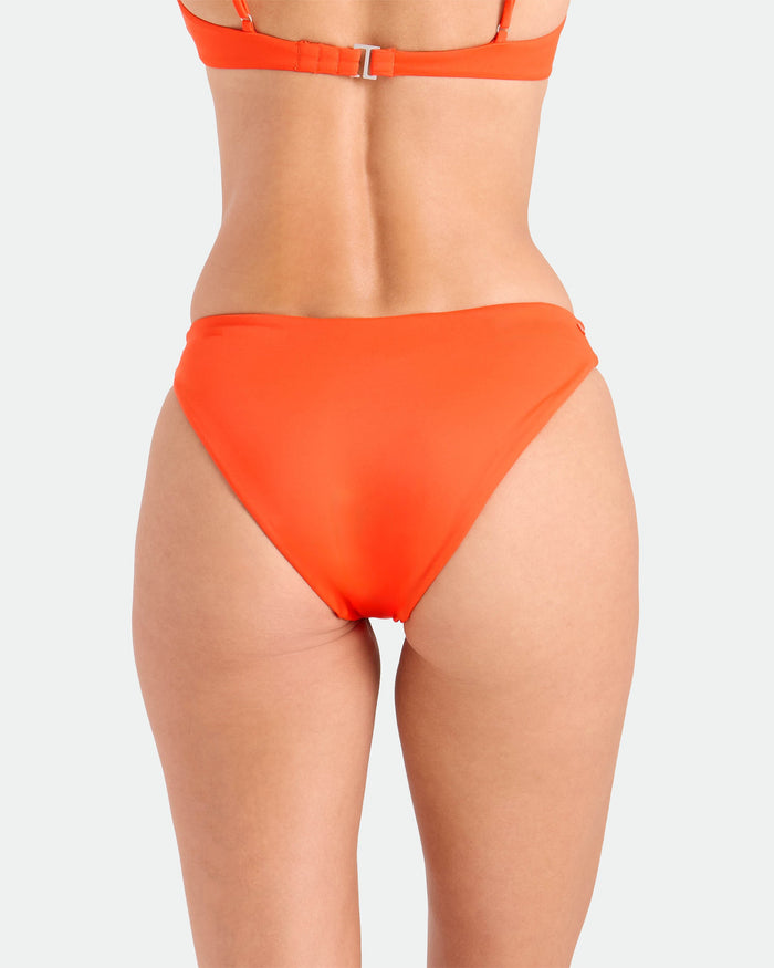 A person is shown from the back, wearing the Volcom Women's Simply Seamless Hipster Full bikini bottom and matching top in bright orange, made from recycled nylon, standing against a plain light background.