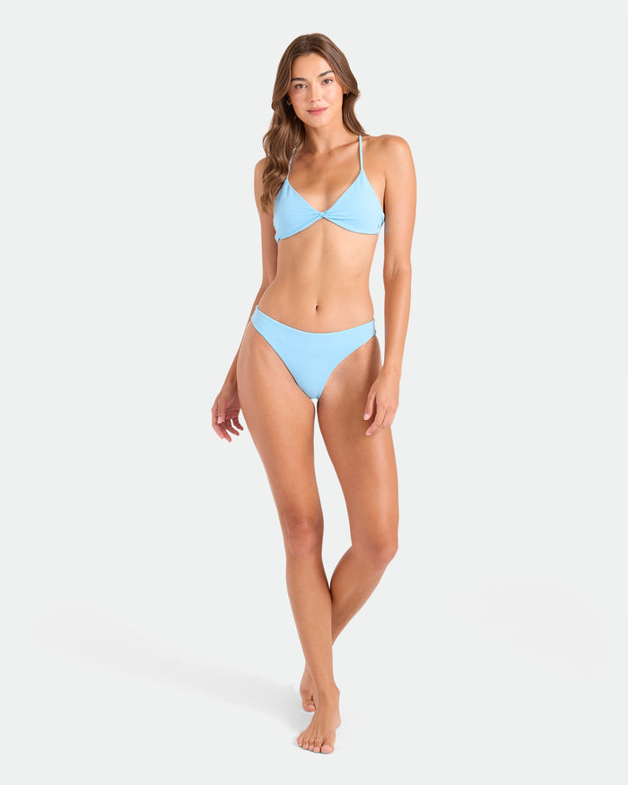 A woman with long brown hair stands barefoot against a white background, modeling the Volcom Women's Simply Seamless Hipster Full light blue bikini made from recycled nylon. She wears a seamless top and full-coverage bottom, exuding relaxed confidence.