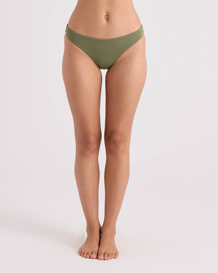 A woman stands barefoot against a plain background, wearing Volcom Women's Simply Seamless Hipster Full bikini bottoms in olive green. Her arms are out of frame, and her body is facing forward, showcasing the comfortable full coverage swimwear design.