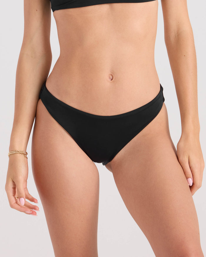 A woman with a neutral skin tone wears the Volcom Women's Simply Seamless Midrise Skimpy black bikini bottom and matching top, made from recycled nylon. Gold bracelet on her left wrist; shown from shoulders to upper thighs.