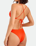 A woman is shown from the back wearing Volcom's Women's Simply Seamless Midrise Skimpy bikini in bright orange, featuring crisscross straps and a tie back. The recycled-nylon set offers cheeky coverage against a plain white background.