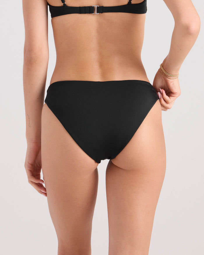A woman is seen from the back wearing the Volcom Women's Simply Seamless Midrise Skimpy bikini bottom and a matching black top made from recycled nylon, with a bracelet on one wrist against a plain light background.