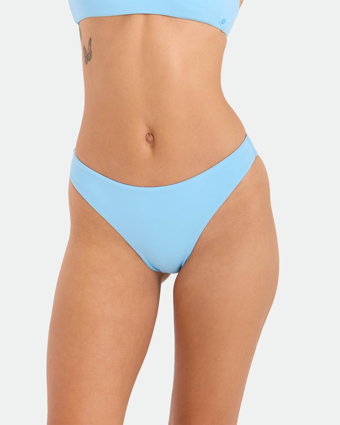 A person wears the Volcom Women's Simply Seamless Midrise Skimpy in light blue, featuring cheeky coverage and recycled nylon, revealing their midsection and a small tattoo near the left ribcage against a plain white background.