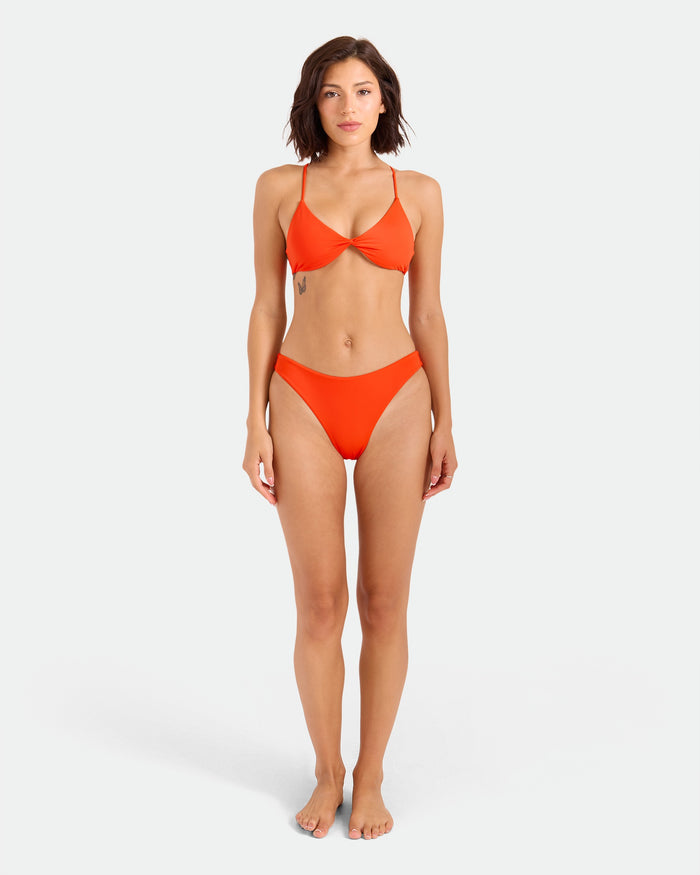 A woman with short brown hair and a neutral expression models the Volcom Women’s Simply Seamless Midrise Skimpy bikini in bright orange, featuring subtle cheeky coverage and recycled nylon, barefoot on a plain light background.