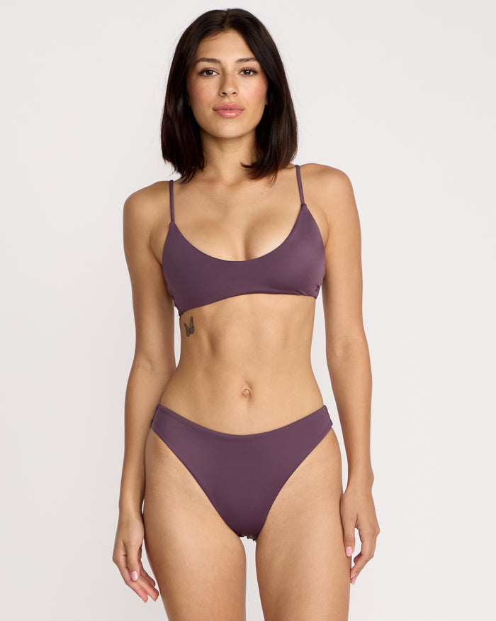 A woman with shoulder-length dark hair and a small butterfly tattoo on her left ribcage poses against a plain background, wearing the mauve Volcom Women's Simply Seamless Skimpy swimwear set. She looks directly at the camera with a neutral expression.