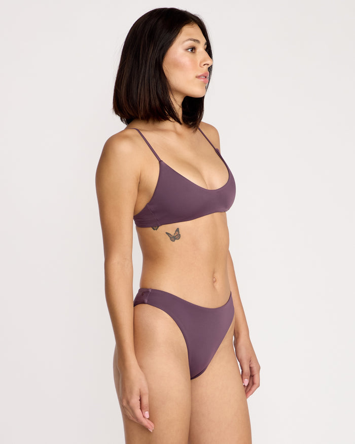 A woman with straight dark hair, wearing the Volcom Women's Simply Seamless Skimpy mauve bikini, stands in profile against a plain white background. A small butterfly tattoo is visible on her left side below her ribs.