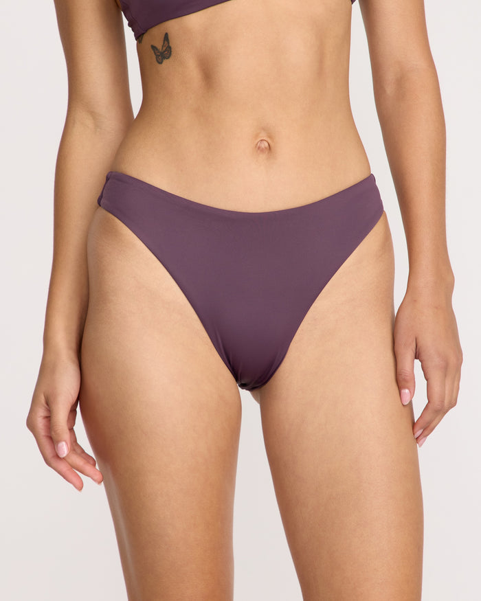 A woman wears the Volcom Women's Simply Seamless Skimpy high-cut purple bikini made from recycled nylon. A small butterfly tattoo is visible below her left ribcage. The background is plain and light-colored.