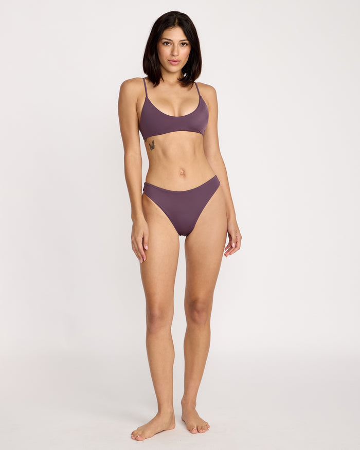 A woman with medium skin tone and dark hair stands barefoot against a plain background, wearing the Volcom Women's Simply Seamless Skimpy bikini in mauve. She looks at the camera with a relaxed expression, one hand by her side.