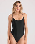 A woman with wavy, light brown hair is wearing the Volcom Women's Simply Seamless Crossback 1Pc swimsuit with thin straps, accessorized with gold jewelry and smiling softly against a plain light background.
