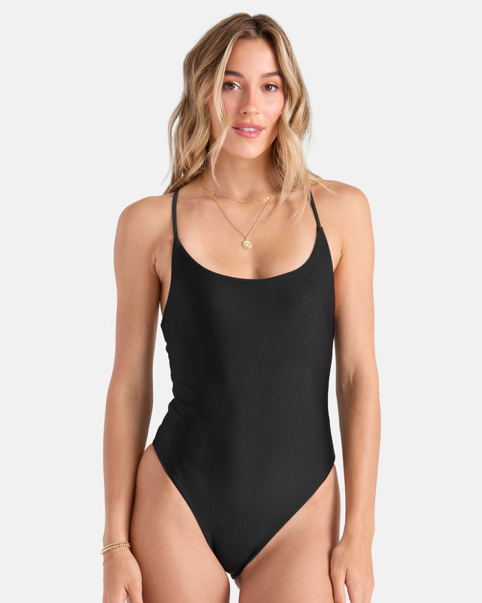A woman with wavy blonde hair wears the Volcom Women's Simply Seamless 1PC Skimpy, a black one-piece made from recycled nylon swimwear. She accessorizes with layered gold necklaces and a bracelet, smiling against a plain light background.