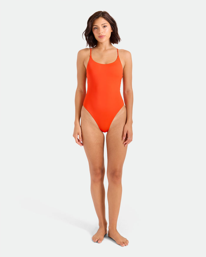 A woman with short dark hair stands barefoot against a plain background, wearing the Volcom Women's Simply Seamless Skimpy One-Piece in bright orange, made from recycled nylon and featuring thin adjustable cross-back straps.