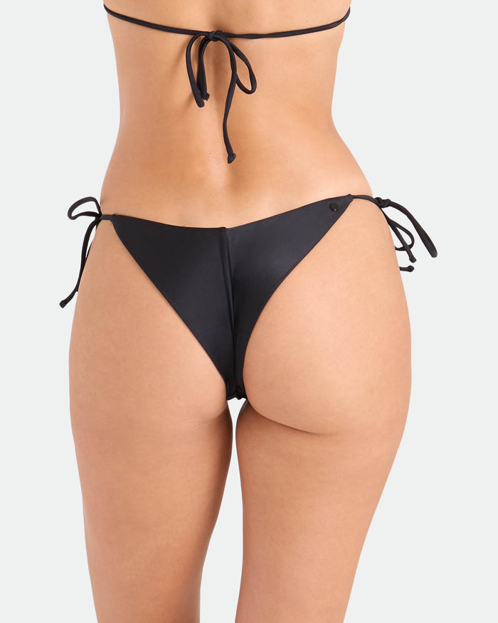 A woman models the Volcom Women's Simply Seamless Tie Side Cheekini Bikini Bottom in black, featuring cheeky coverage, viewed from the back against a plain white background.