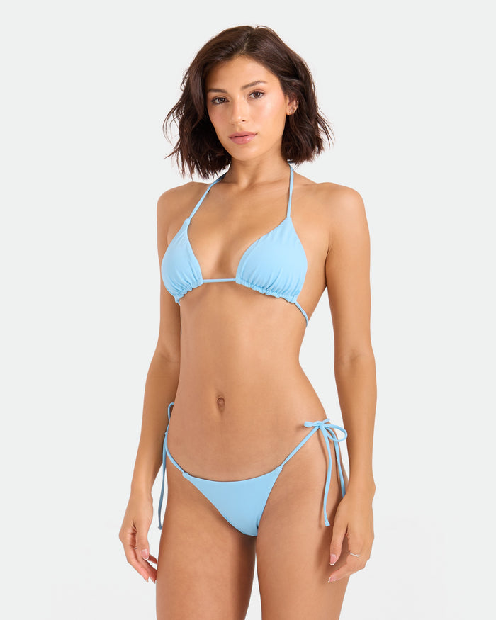 A woman with short brown hair wears the Volcom Women's Simply Seamless Tie Side Cheekini Bikini Bottom, made from recycled nylon, standing against a plain white background and looking at the camera with a neutral expression.