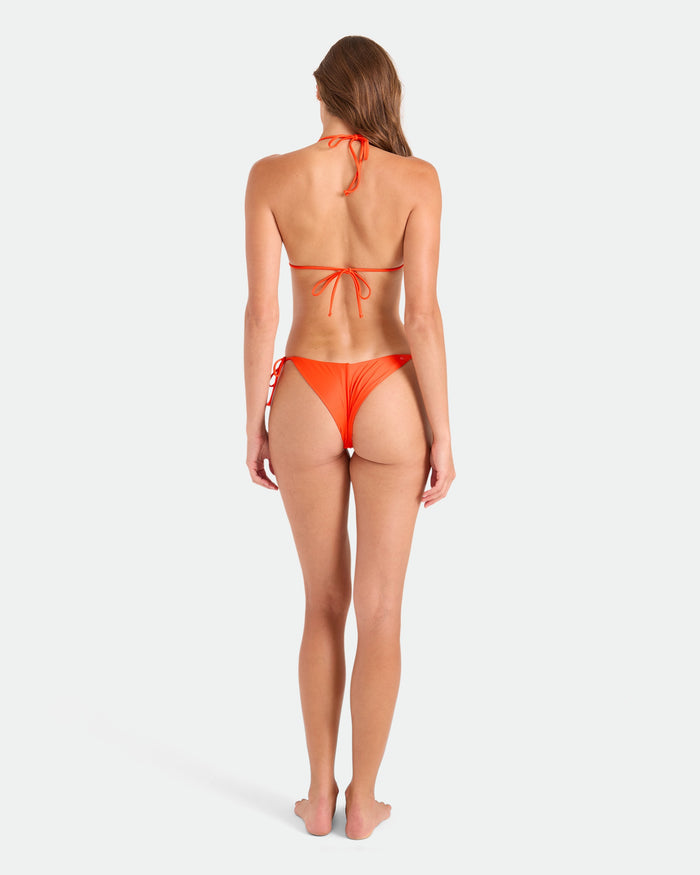 A woman with long brown hair stands barefoot facing away, wearing the Volcom Women's Simply Seamless Tie Side Cheekini Bikini Bottom in orange, made from recycled nylon, against a plain white background.