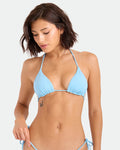 A woman with short brown hair models the Volcom Women's Simply Seamless Triangle bikini top in light blue, featuring adjustable tie straps. She looks down with one hand on her hip, showing a small butterfly tattoo. The background is plain and light.