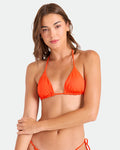 A woman with straight brown hair models the Volcom Women's Simply Seamless Triangle bikini top and matching bottoms in orange, standing against a plain light background with one arm crossed, gazing at the camera with a relaxed look.