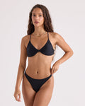 A woman with long wavy brown hair wears the Volcom Women's Simply Seamless Twist Bralette and gold jewelry, posing confidently against a plain light background with one hand on her hip.