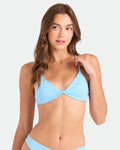 A woman with long brown hair wears the Volcom Women's Simply Seamless Twist Bralette in light blue, made from recycled nylon, gazing to the side with a relaxed expression against a plain, light background.