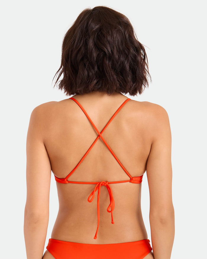 A woman with short, wavy brown hair is shown from the back wearing the Volcom Women's Simply Seamless Twist Bralette in orange, featuring crisscrossed straps and a tie at the back against a plain, light-colored background.