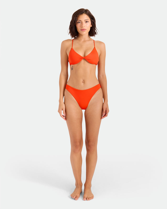 A woman with shoulder-length brown hair stands barefoot against a white background, wearing the Volcom Women's Simply Seamless Twist Bralette in orange with matching recycled nylon bottoms, and has a relaxed, neutral expression.
