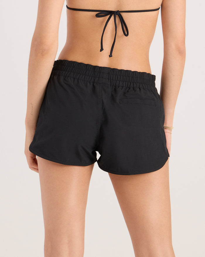 A woman is seen from the back in a black bikini top and Volcom Women's Simply Solid Boardshorts, made with recycled polyester and a 4-way stretch elastic waistband, set against a plain neutral background.
