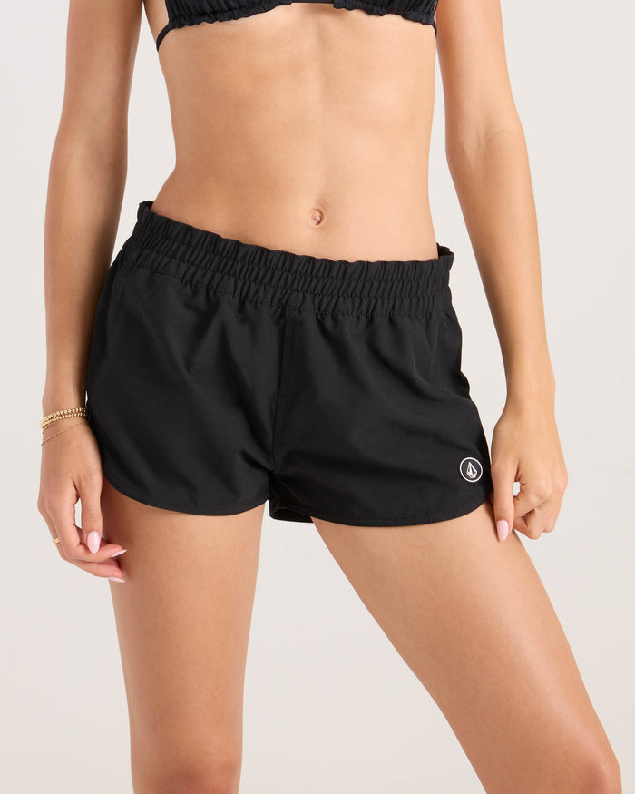 A woman wears the Volcom Women's Simply Solid Boardshort—black, 4-way stretch, elastic waistband, small left leg logo—paired with a matching top. Her left hand is on her thigh and she wears a gold bracelet on her right wrist.