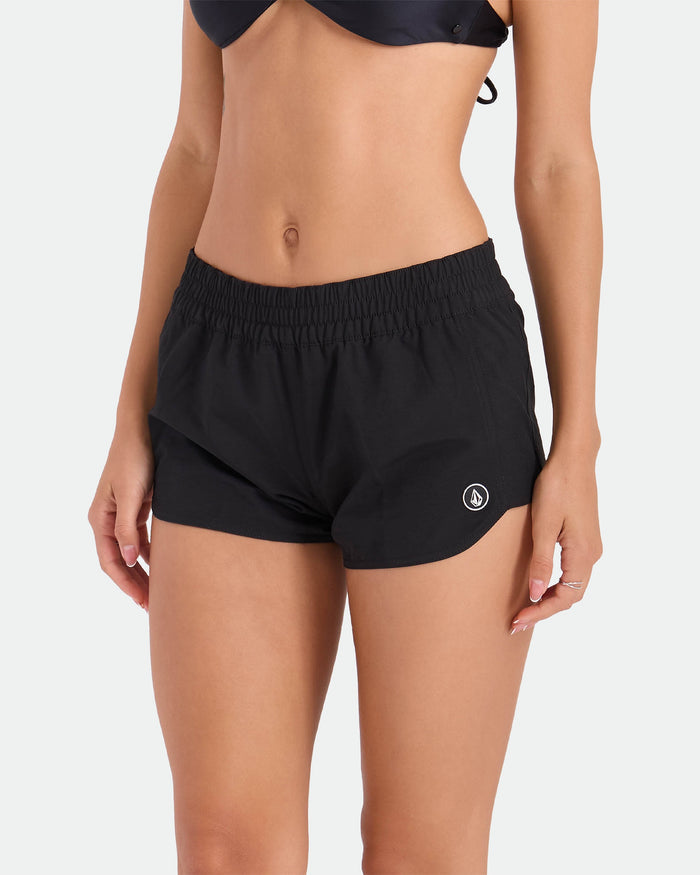 A woman models the Volcom Women's Simply Solid Boardshort, featuring an elastic waistband and a small white logo on the left leg. She pairs it with a black bikini top and stands against a plain light background. Her face is not visible.