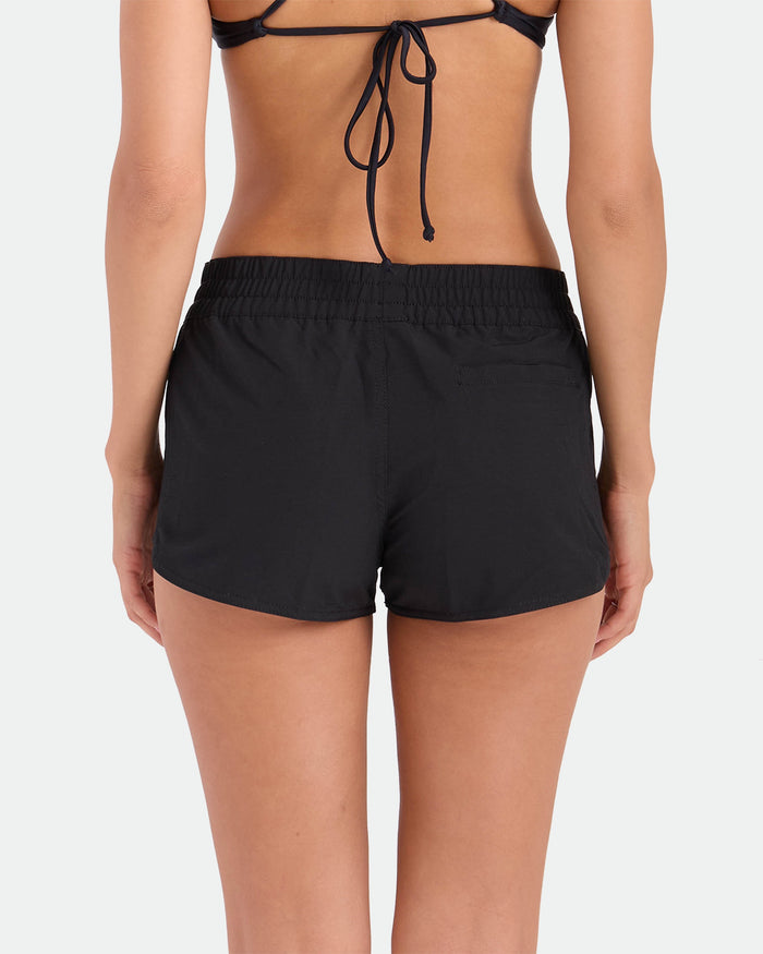 A woman is seen from the back in a black bikini top and Volcom Women's Simply Solid Boardshorts with an elastic waistband, standing against a plain white background.