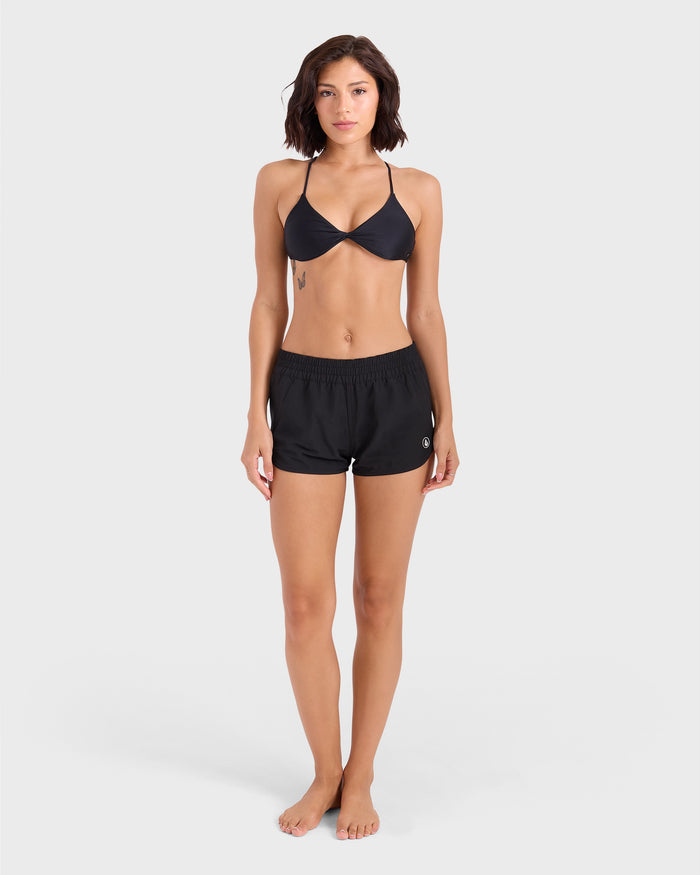 A woman stands barefoot against a plain light background, facing forward with arms relaxed by her sides, wearing a black bikini top and Volcom Women's Simply Solid Boardshort made from recycled polyester.