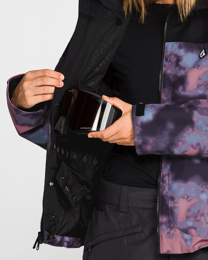 A person clad in the Volcom Women’s Steele Insulated Jacket in a purple tie-dye pattern opens an inner pocket to reveal a black and white device inside, with only their torso and hands visible against a plain background.