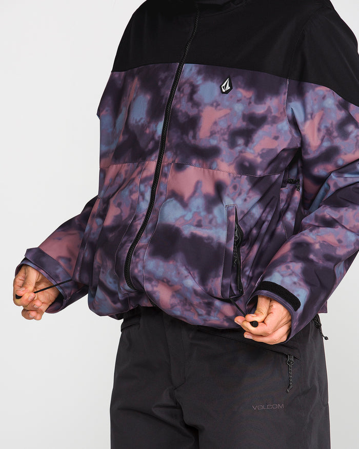 A person wearing the Volcom Women’s Steele Insulated Jacket in a purple and black pattern with a zipper, paired with black pants, stands against a plain light background; only the lower face and torso are shown.