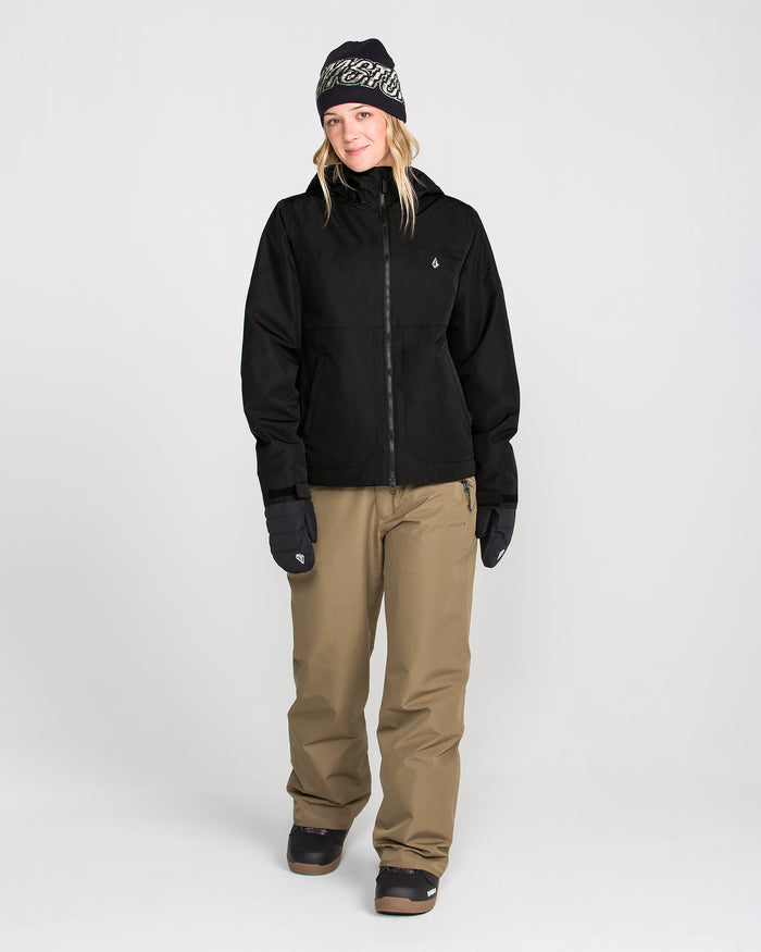 Wearing the Volcom Women’s Steele Insulated Jacket, a person pairs it with tan snow pants, black gloves, a black beanie, and snow boots as they stand against a plain white background, smiling slightly.