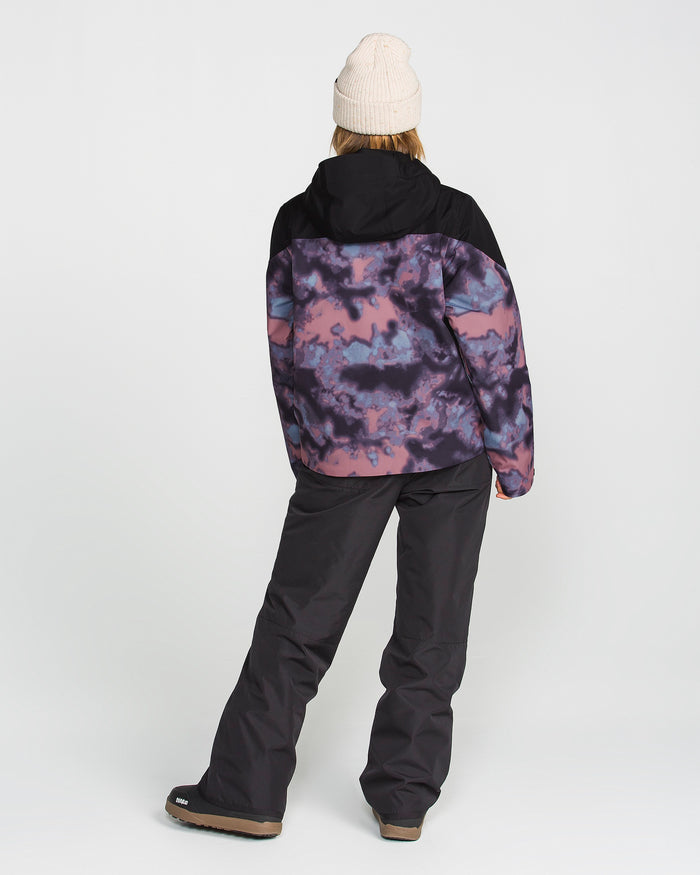 Wearing the Volcom Women’s Steele Insulated Jacket—styled in a purple and black cloud pattern with eco-friendly recycled insulation—a woman stands facing away against a plain white background. She pairs the hooded, waterproof jacket with simple black pants and a light knit hat for a modern, sustainable look.