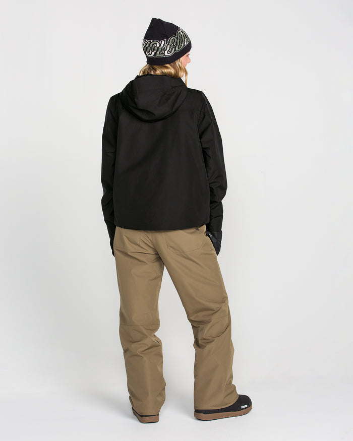 A person stands facing away, wearing the Volcom Women’s Steele Insulated Jacket in black—featuring recycled insulation—along with tan pants, black gloves, black boots, and a black knit hat with white lettering, against a plain white background.