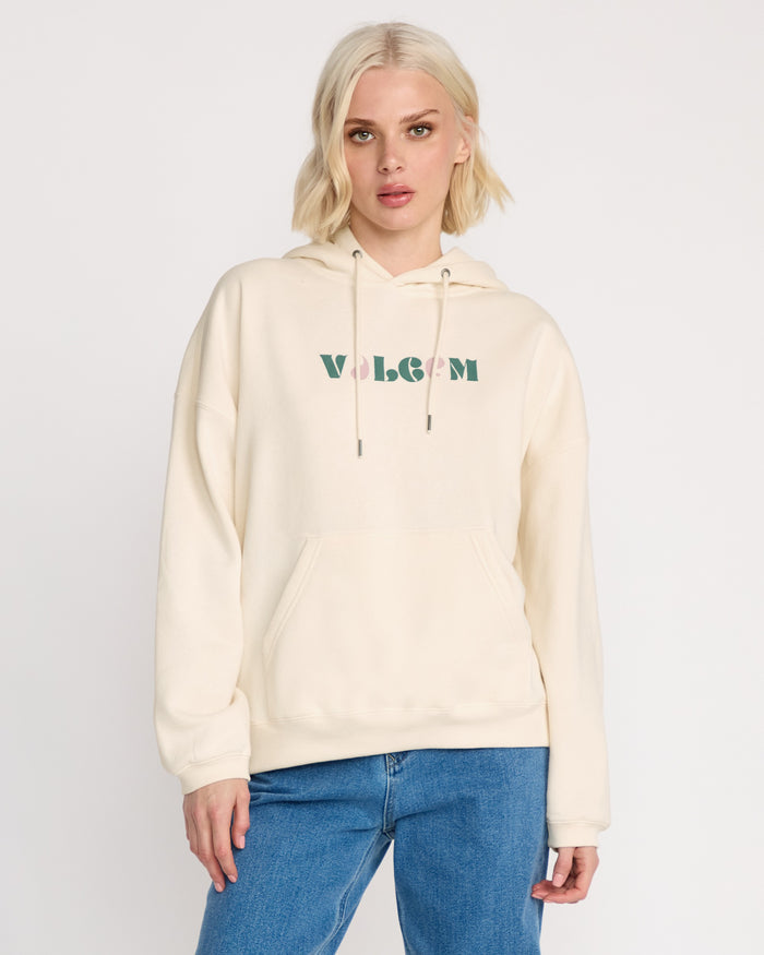 A woman with short blonde hair models the Volcom Women's Stone Art Up Hoodie, a cream-colored oversized style with green and pink VOLCOM logo, paired with blue jeans against a white background—ideal for relaxed streetwear looks.