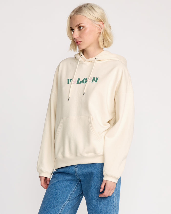 A woman with short blonde hair models the Volcom Women's Stone Art Up Hoodie in cream-colored brushed fleece and blue jeans, standing against a plain light background with a neutral expression for a modern streetwear look.