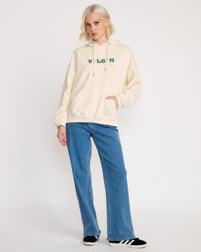 A woman with short blonde hair wears the Volcom Women's Stone Art Up Hoodie, cream-colored and oversized, with blue jeans and black-and-white sneakers, showing off streetwear style against a plain white background.