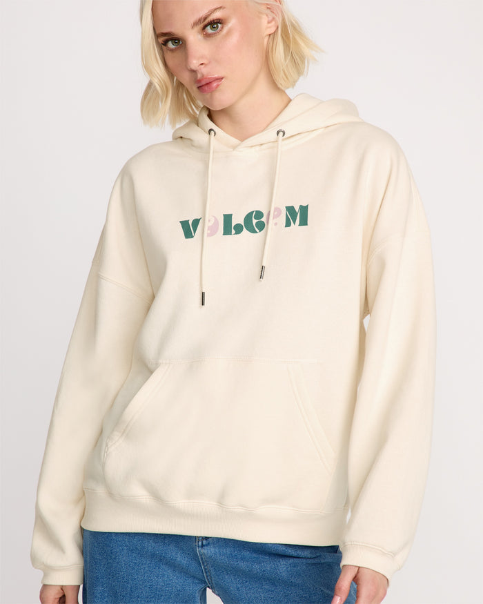 A woman with short blonde hair wears the oversized cream-colored Volcom Women's Stone Art Up Hoodie with green "VOLCOM" lettering and blue jeans, standing against a plain light background.