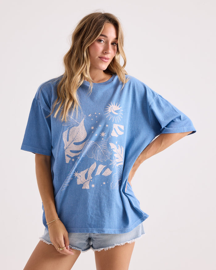 A woman with long wavy hair wears the Volcom Women's Stone Breaker Tee—a loose-fitting, 100% cotton shirt featuring blue fabric with white graphic designs—paired with light denim shorts. She stands against a plain light background, one hand resting on her hip.